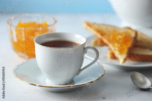 A cup of black tea and a saucer. Next to the plate with toast and a vase with orange jam. In the frame, a teaspoon and a teapot. Light background. Close-up. Macro photography.