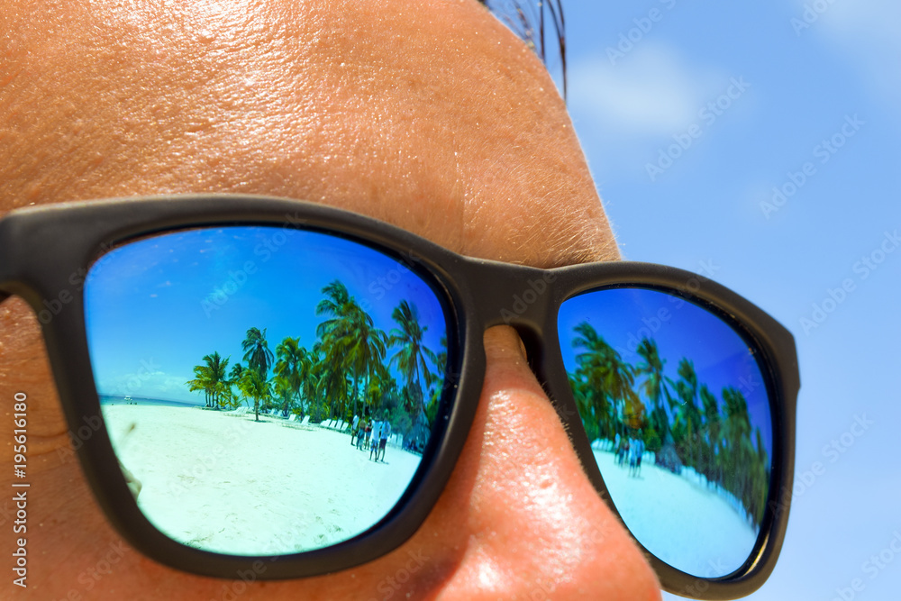 Reflection of white beach with palms in the man's sunglasses, man at the  beach with sunglasses Stock Photo