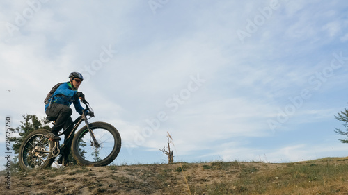 Fototapeta Naklejka Na Ścianę i Meble -  Fat bike also called fatbike or fat-tire bike in summer driving through the hills. The guy is riding a bike along the sand and grass high in the mountains. He performs some tricks and runs dangerously