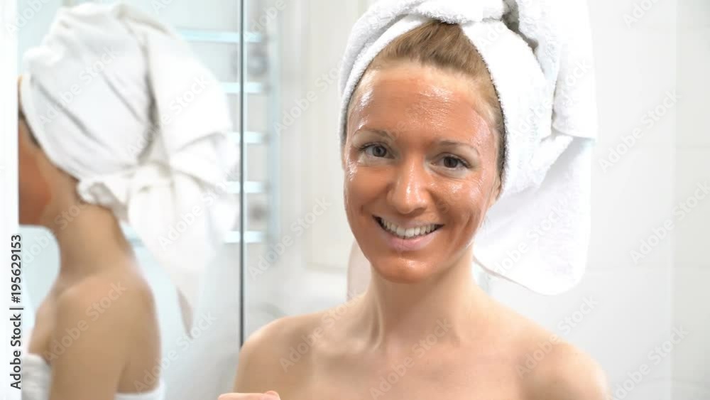 a young woman with a white towel put on her face a brown moisturizing mask and smiling at the camera a young woman with a white towel put on her face a brown moisturizing mask and smiling at the camera