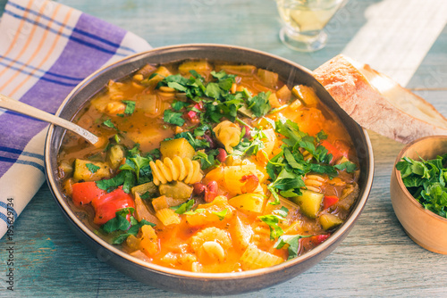 Traditional minestrone soup with pasta and chopped green parsley in a plate on a rustic wooden table close-up with a napkin, sunny day - top view, copy space for a recipe