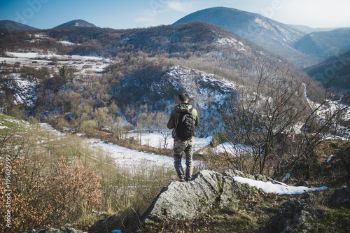 Young man standing on top of cliff in winter mountains holding a camera and enjoying view.