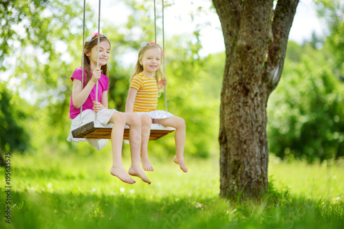 Two cute little sisters having fun on a swing together in beautiful summer garden on warm and sunny day outdoors © MNStudio