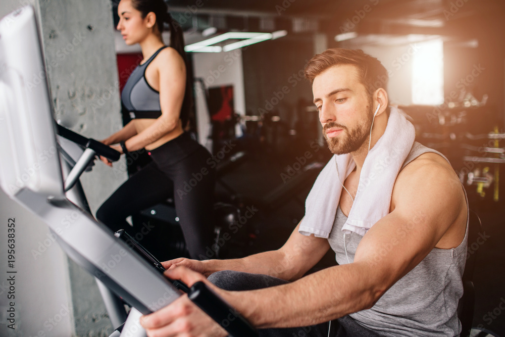 Close up of a guy wokring on the exercise bike and his girfriend doing the same thing further down. Both of them are serious and concentrated.