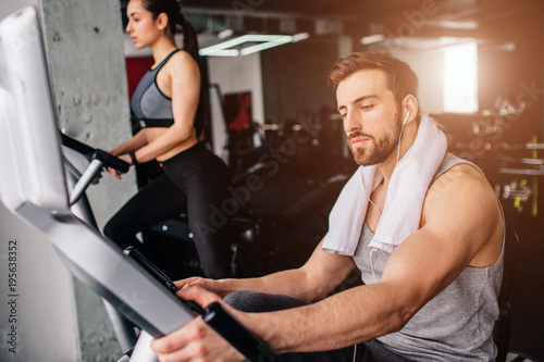 Close up of a guy wokring on the exercise bike and his girfriend doing the same thing further down. Both of them are serious and concentrated.