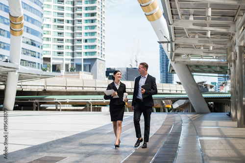 Asian business woman wear suit holding document file on hand and walking together with  Caucasian businessman in the city and talk about business future.