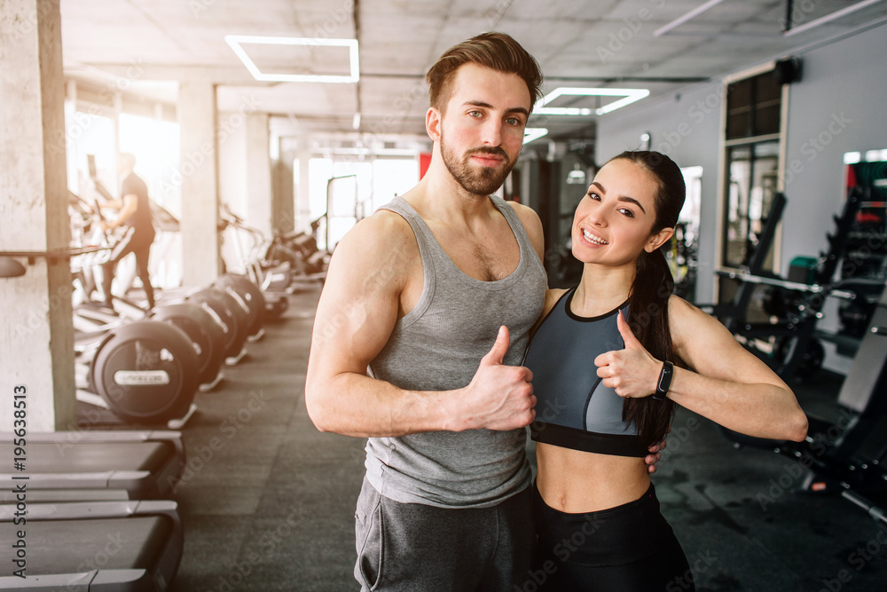 A picture of a couple standing in the sport club's training room together and showing their big thumb's up. They are happy to exsercise in this fitness club. Young couple is smiling.