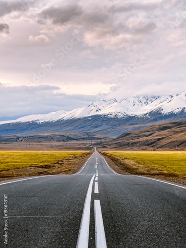 Inspirational travel and adventure photography. Straight paved road with white surface markings goes through the snow capped mountains of Altai Republic, Russia just before the sunset.