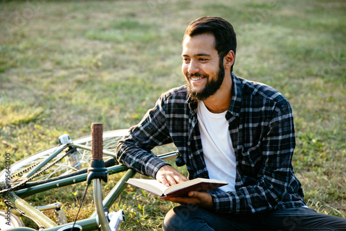 Handsome happy bearded man reading interesting book, spending time with pleasure while sitting on the grass near the bike.