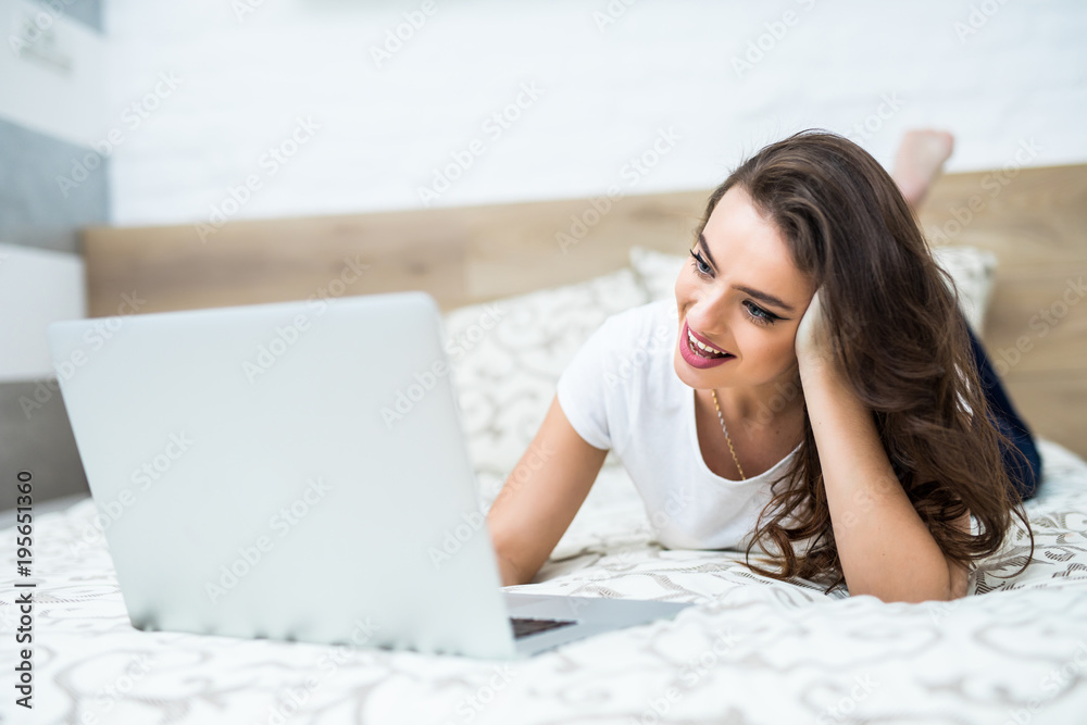 Happy young girl on the phone lying in bed while using laptop Happy young girl on the phone lying in bed while using laptop