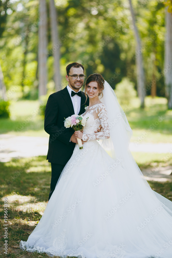 Beautiful wedding photosession. The groom in a black suit and his bride in a white lace dress with a long plume, veil and bouquet smile and embrace in a large green garden on weathery sunny day