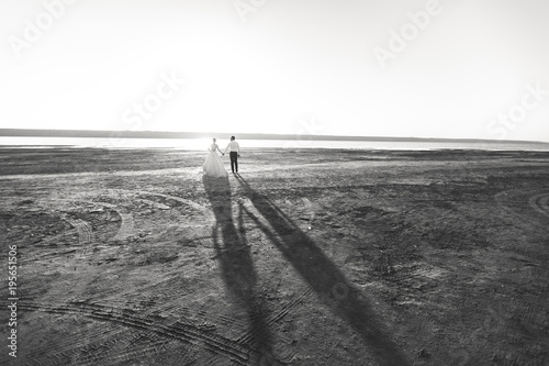 Beautiful wedding photosession. Handsome unshaved groom in a black trousers and young cute bride in white lace pattern dress with exquisite hairstyle on walk along the coastline near the sea sunset