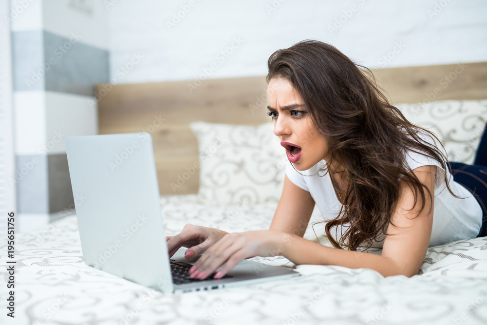 Surprised young woman lying on the bed and looking at laptop Surprised young woman lying on the bed and looking at laptop