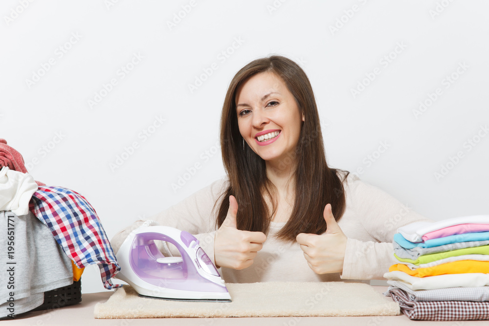 Young smiling housewife in light clothes with colorful family clothing on ironing board with iron. Woman showing thumbs up isolated on white background. Housekeeping concept. Copy space advertisement. Young smiling housewife in light clothes with colorful family clothing on ironing board with iron. Woman showing thumbs up isolated on white background. Housekeeping concept. Copy space advertisement.