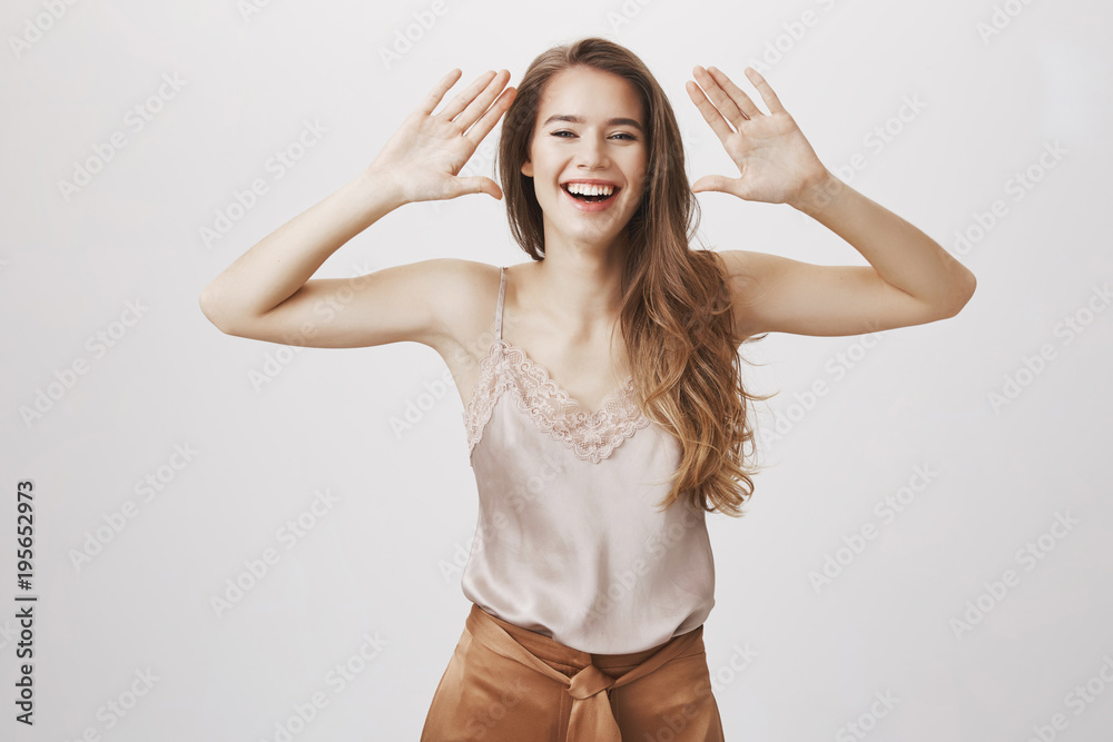 Beautiful emotions make people shine. Portrait of attractive european female with beautiful white smile holding palms near face and laughing, enjoying great day at party, standing over gray wall