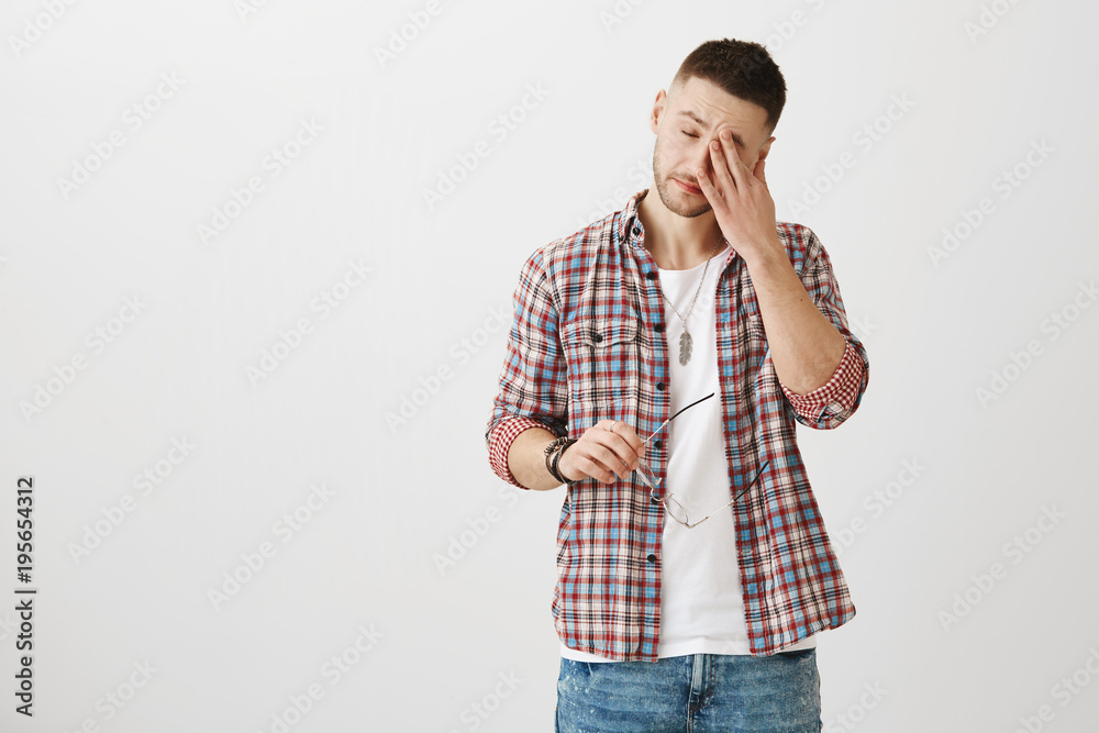 Portrait of tired ordinary european guy with bristle, holding glasses while rubbing eyes, being exhausted and wanting to get rest against gray background. Man spent too much time near laptop Portrait of tired ordinary european guy with bristle, holding glasses while rubbing eyes, being exhausted and wanting to get rest against gray background. Man spent too much time near laptop