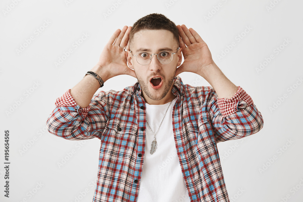Where did you hear that from. Shocked and interested young handsome male with bristle in trendy eyewear holding palms near ears to listen carefully, standing with open mouth while gossiping Where did you hear that from. Shocked and interested young handsome male with bristle in trendy eyewear holding palms near ears to listen carefully, standing with open mouth while gossiping