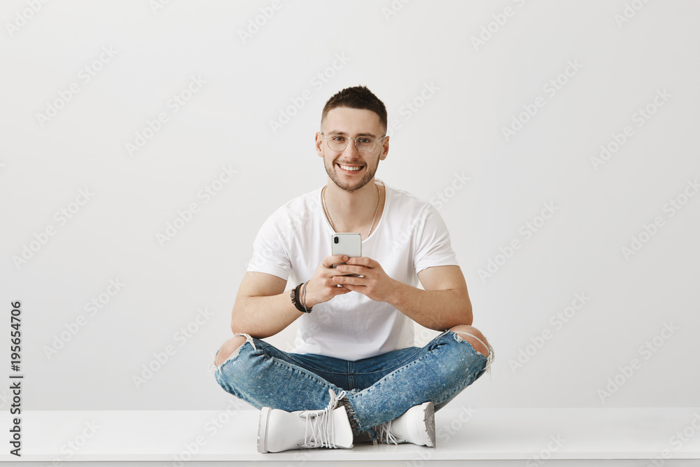 Attractive young caucasian male student of university sitting on floor with crossed legs in transparent glasses, holding smartphone while smiling at camera over gray background. Attractive young caucasian male student of university sitting on floor with crossed legs in transparent glasses, holding smartphone while smiling at camera over gray background.
