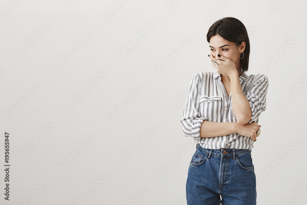 Naklejka premium Attractive caucasian woman with short dark hair in stylish casual clothes mocking and chuckling, covering mouth, trying to stop laughing, being in playful mood. Girl sees how her boyfriend slipped