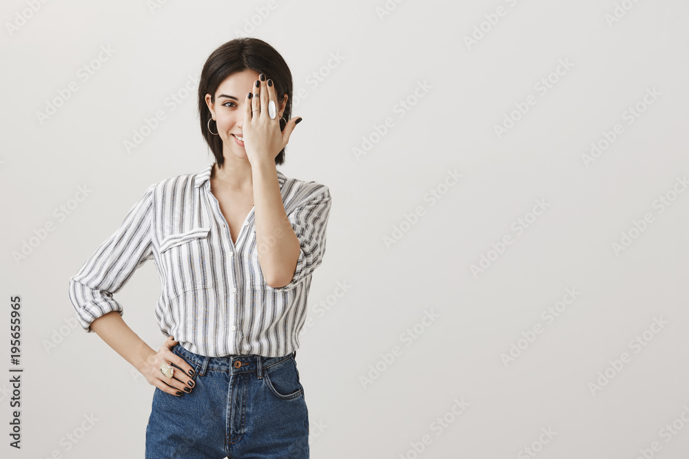Jewellery is best friend of women. Attractive european girl with short dark hair in stylish clothes smiling and holding hand on hip while covering half of face with palm, showing trendy ring Jewellery is best friend of women. Attractive european girl with short dark hair in stylish clothes smiling and holding hand on hip while covering half of face with palm, showing trendy ring