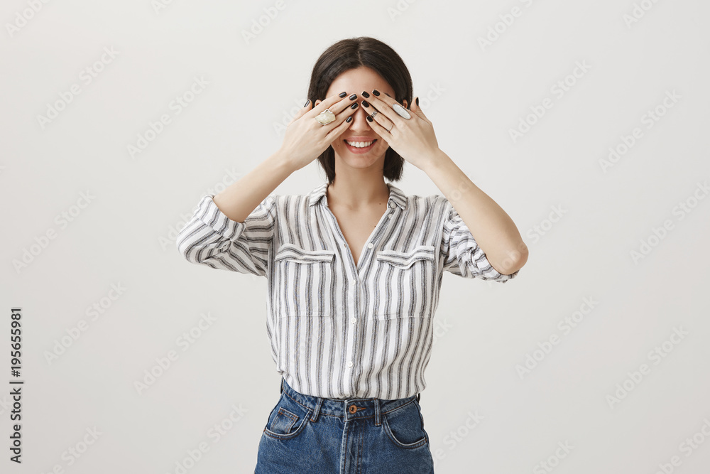 Promise not to peep. Studio shot of attractive playful woman in stylish clothes and in good mood smiling broadly while covering eye with hands, showing trendy rings, standing over gray background Promise not to peep. Studio shot of attractive playful woman in stylish clothes and in good mood smiling broadly while covering eye with hands, showing trendy rings, standing over gray background