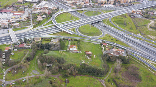 Aerial view of a series of ramps of the Rome motorway junction. Many cars, motorcycles and trucks drive on the road. Around there are grass, meadows and green trees.