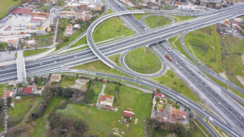 Aerial view of a series of ramps of the Rome motorway junction. Many cars, motorcycles and trucks drive on the road. Around there are grass, meadows and green trees.