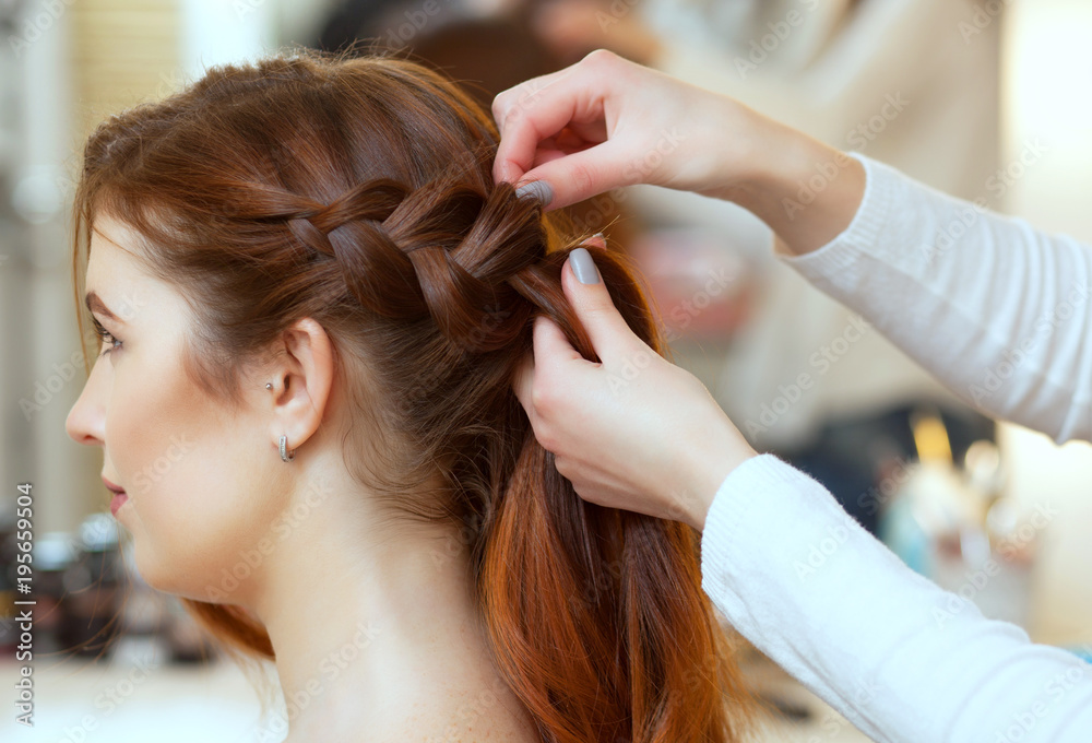 Obraz premium Beautiful, red-haired girl with long hair, hairdresser weaves a French braid, in a beauty salon. Professional hair care and creating hairstyles.
