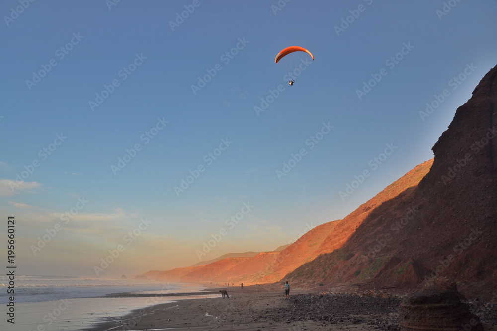 The paraglider flies over the coast at sunset. Red rocky coast of Legzira beach, Morocco.