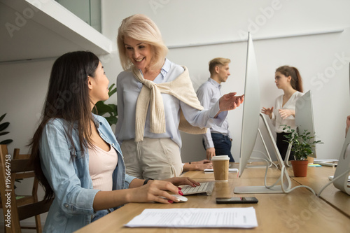 Smiling friendly senior female executive talking to asian employee working on pc, happy aged leader telling young coworker good news, excited older mentor supporting achievements of intern in office