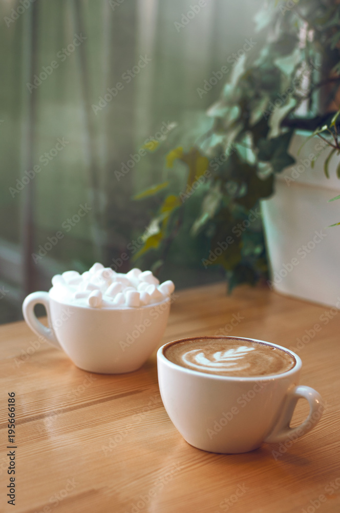 Two cups of cappuccino with latte art and marshmallow on wooden background. Beautiful foam, greenery ceramic cups, stylish toning, place for text.