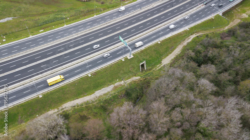 Aerial view of a stretch of highway in the Italian countryside. The road is divided into many lanes in each direction. There are cars and trucks on the street.Around the road there are trees and grass