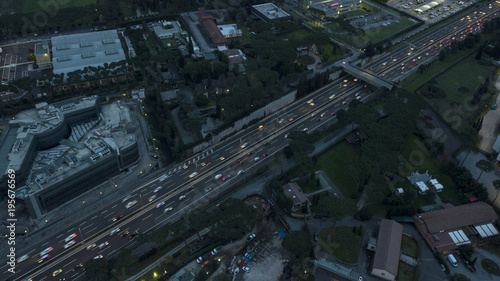 Aerial night view of a highway junction in Rome, Italy. The street crosses an industrial area. On the road, many cars drive at high speed with the headlights on. Around there are grass and trees.