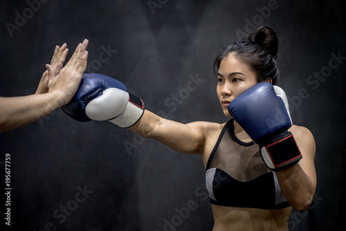 Young Asian woman boxer with blue boxing gloves punching to her trainer’s hand in training gym, Martial arts on black background © zephyr_p