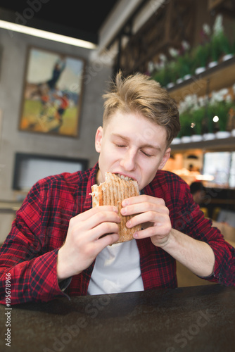 A young man bites a panini sandwich in a cafe. The student is sitting in a cozy cafe and dining with a sandwich