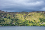 Ullapool, Scotland - June 8, 2012: Hills on west shore of Loch Brrom near Ullapool is green and brown with small white house hidden in green, all under dark cloudscape darkening the water.