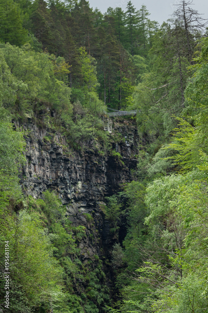 Fototapeta premium CBraemore, Scotland - June 8, 2012: Corrieshalloch Gorge, a deep cut in landscape with forested vertical slopes. Suspension bridge over chasm. Focus on black rocks and abundance of green trees.