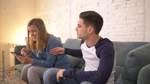 panning shot of young woman using social media on mobile phone ignoring her boyfriend at home sofa couch with the guy frustrated upset bored ignored by his girfriend in internet addict photo