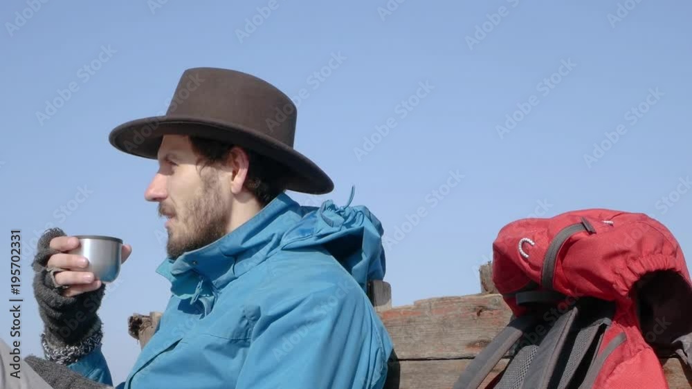 00:03 | 00:12 1× Young bearded hiker in blue jacket and hat drink coffee or tea on the beach, old wooden boat background 00:03 | 00:12 1× Young bearded hiker in blue jacket and hat drink coffee or tea on the beach, old wooden boat background