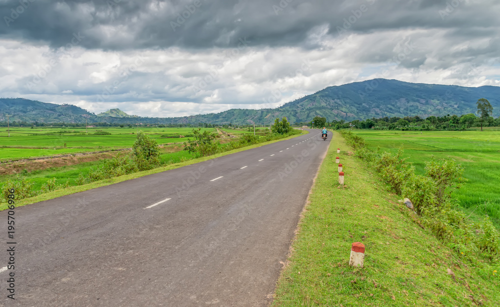 asphalt road, among the fields, with people riding a moped towards the mountains, against the background of clouds, sky and green vegetation