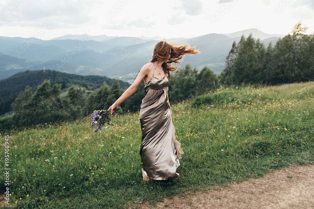 A beautiful girl in a dress and a bouquet of flowers in her hands, on a green glade dancing among the peaks of the mountains
