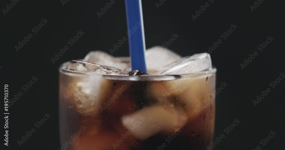 Slow motion closeup put blue straw into cola with ice cubes in glass ove black background