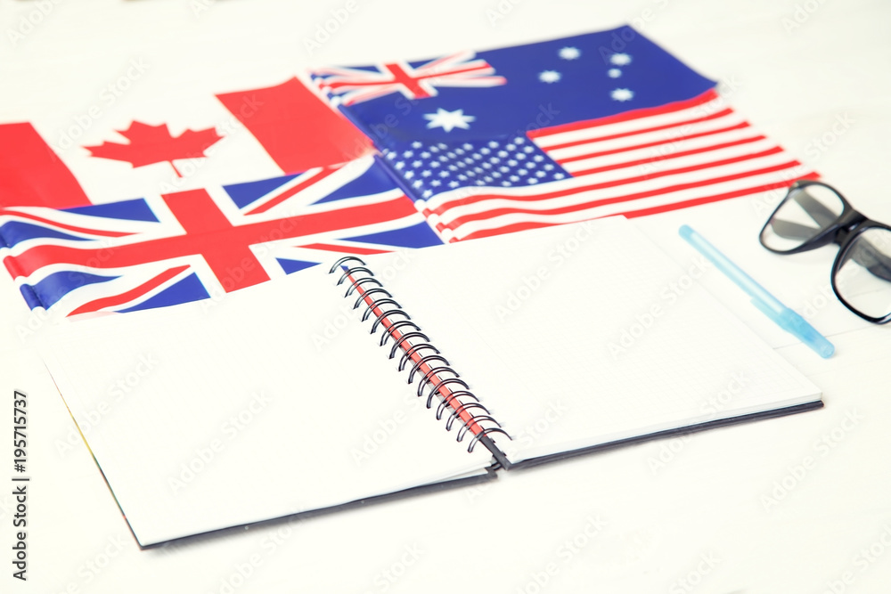 Education concept. English language learning. English speaking countries.  Notebook, textbook, blue pen, glasses, four flags on a wooden table. Photos  | Adobe Stock