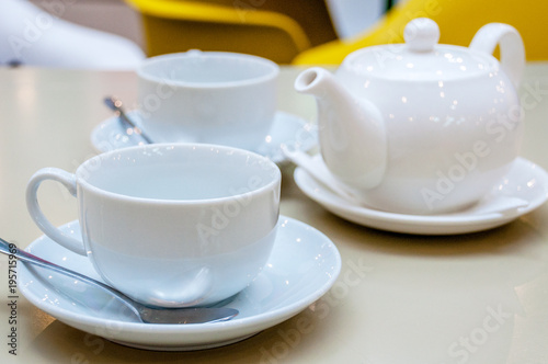 Two white ceramic tea mugs with shiny spoons and saucers and a teapot stand on the table in a cafe