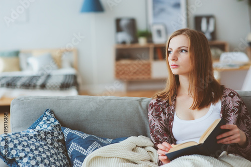 indoor portrait of young thoughtful woman reaxing at home in weekend with interesting book, sitting on cozy couch in modern interior photo