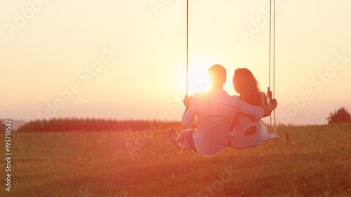 LENS FLARE: Happy couple swaying on a swing embraced on sunlit summer evening.
