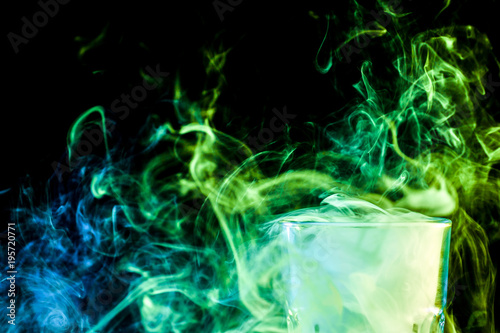 A close-up transparent glass filled with a wig from a green vape smokes and stands on a black isolated background. Glass bongs for smoking soft focus.