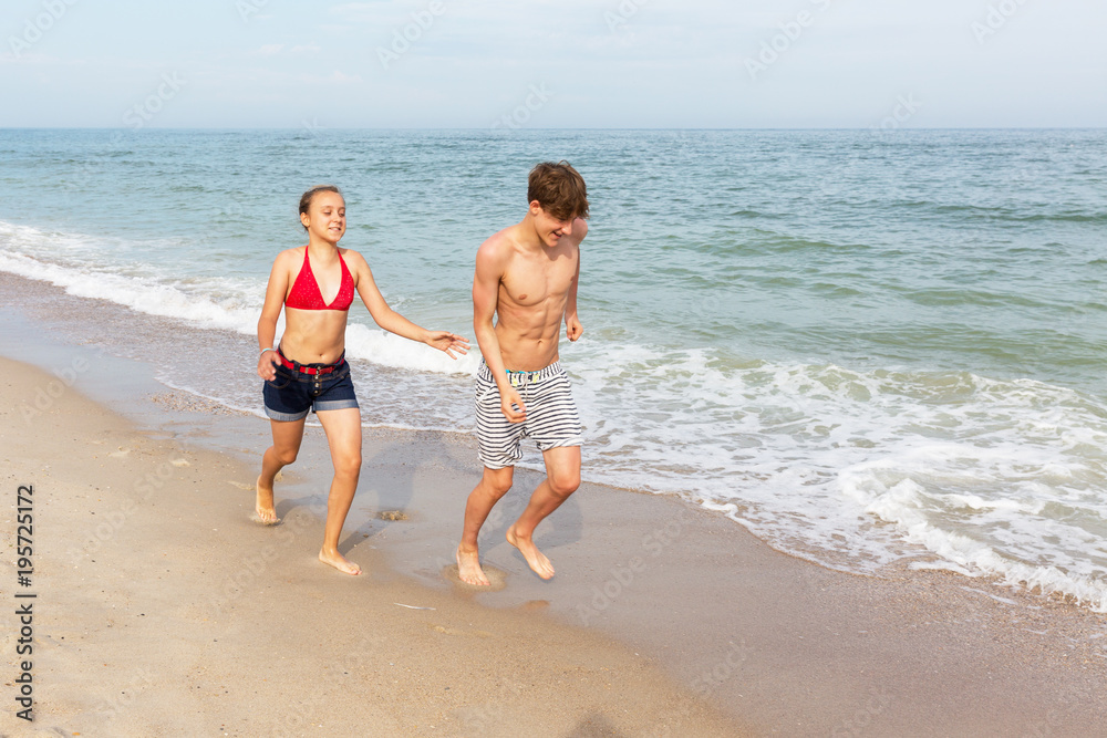 Two teenagers: a girl and a boy with blond hair, dressed in a swimsuit run, laugh and play in the sea beach.