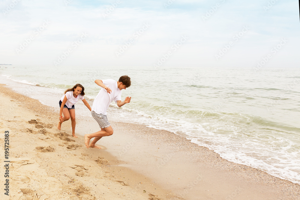 Two teenagers: a girl and a boy with blond hair, dressed in white T-shirts they run, laugh and play in the sea beach.
