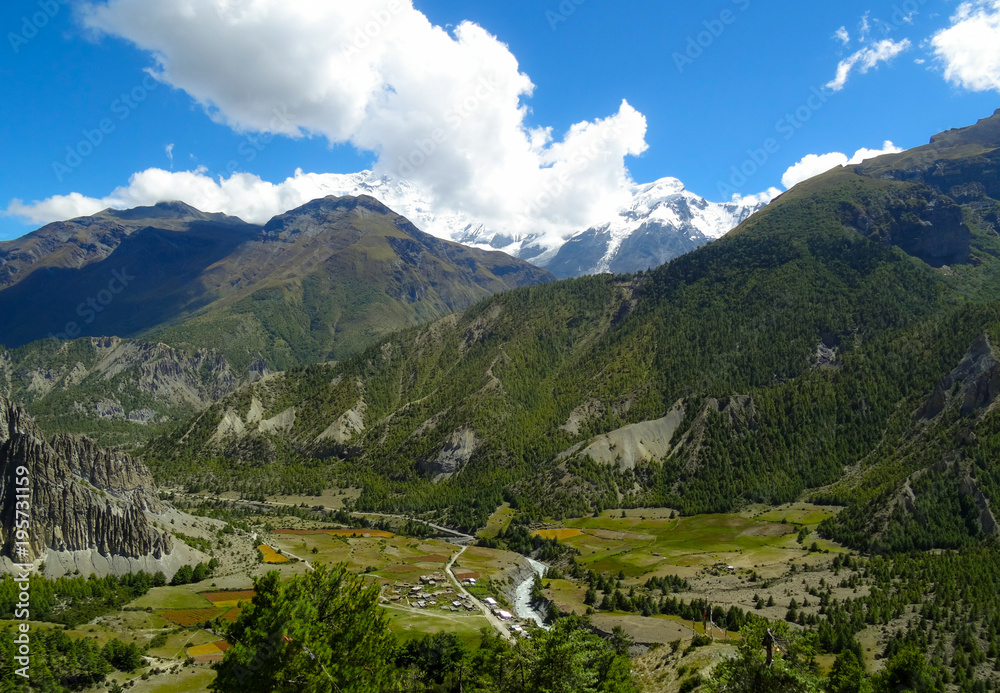 Fototapeta premium A village beside a river below green sided mountains and snow covered mountain tops